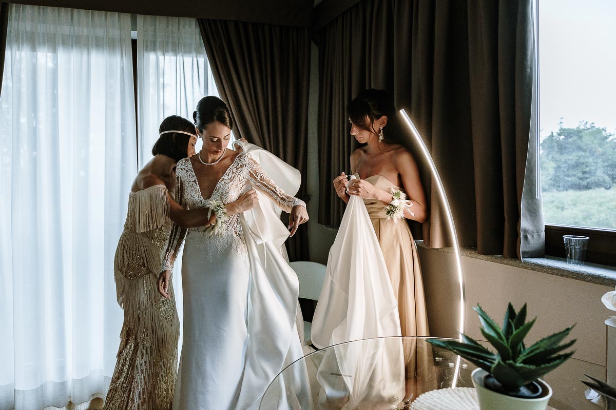 Fotografo Matrimonio Lago di Viverone, Tenuta Variselle. Preparazione della sposa. Sorelle aiutano la sposa ad indossare la sopragonna del vestito.