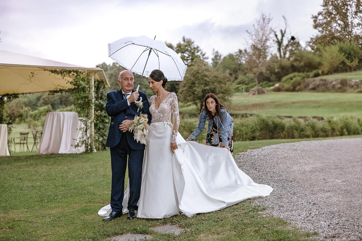 Fotografo Matrimonio Lago di Viverone, Tenuta Variselle. Cerimonia religiosa secondo il rito della Chiesa di Antiochia. Sposa con il padre sotto l'ombrello. La wpsistema l'abito della sposa.