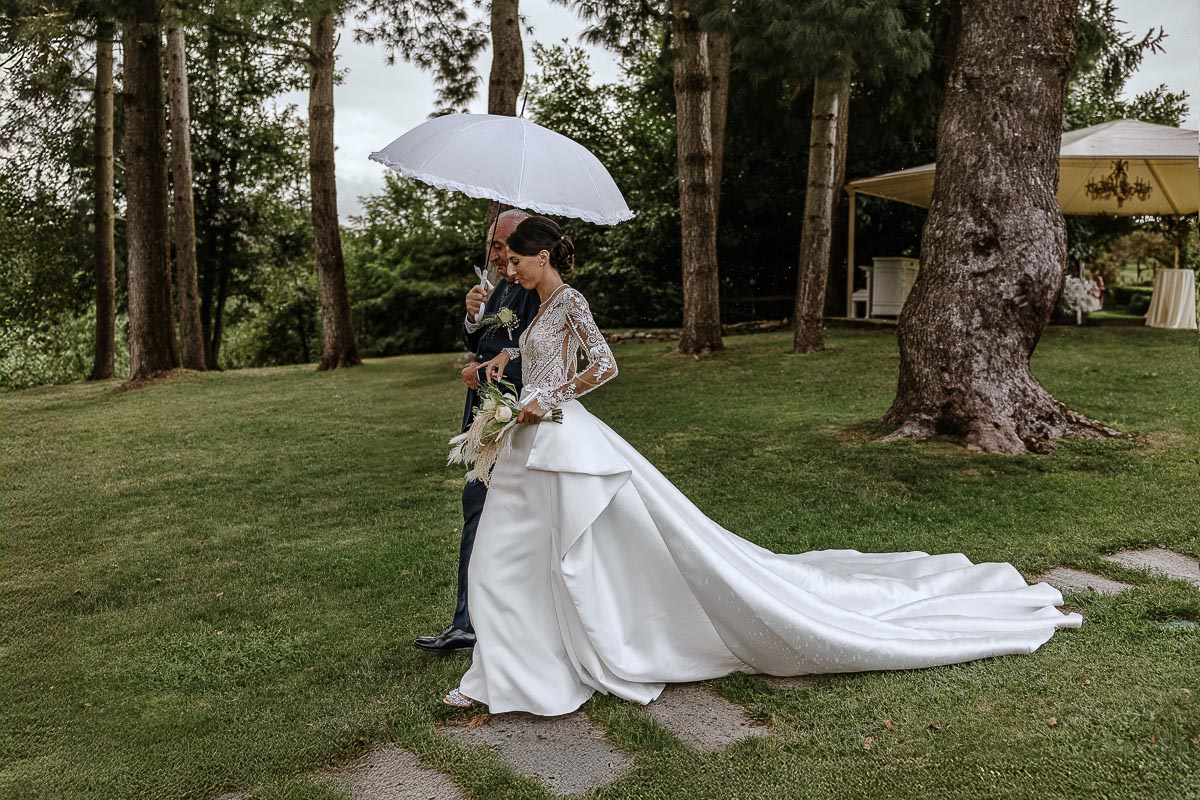 Fotografo Matrimonio Lago di Viverone, Tenuta Variselle. Cerimonia religiosa secondo il rito della Chiesa di Antiochia. Sposa al braccio del padre sotto l'ombrello.