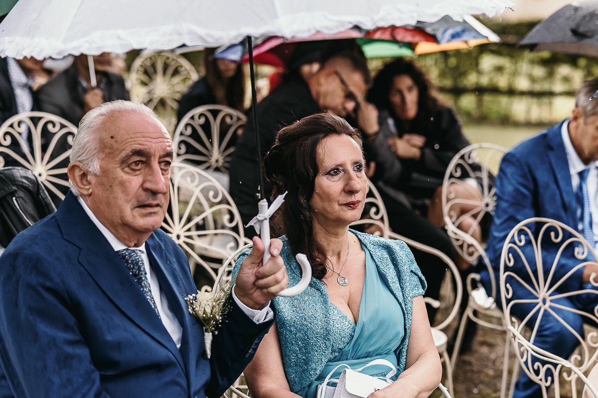 Fotografo Matrimonio Lago di Viverone, Tenuta Variselle. Cerimonia religiosa secondo il rito della Chiesa di Antiochia. genitori della sposa sotto l'ombrello.