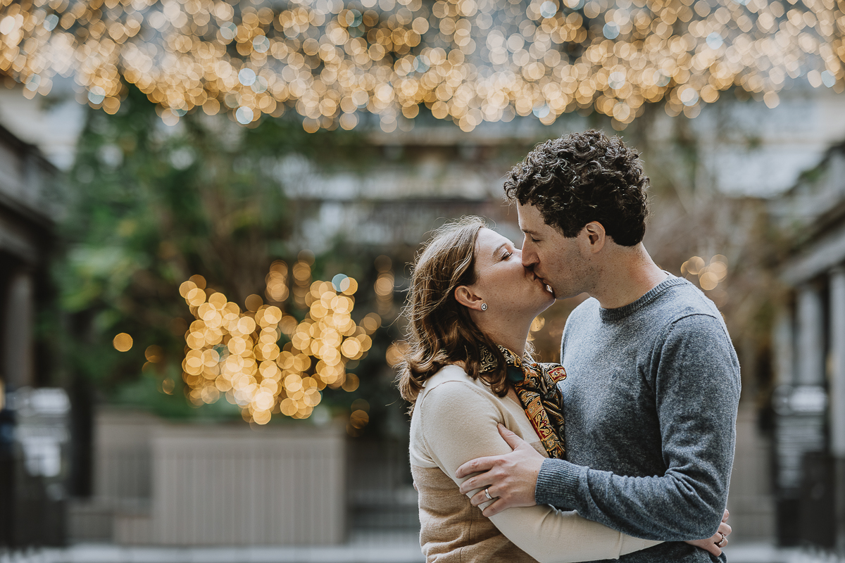 Servizio fotografico per famiglia americana in vacanza a Torino. Papà e mamma si baciano nel bosco urbano delle Gallerie d'Italia nel cortile della Banca san Paolo in piazza San Carlo con le luci di Natale sfocate