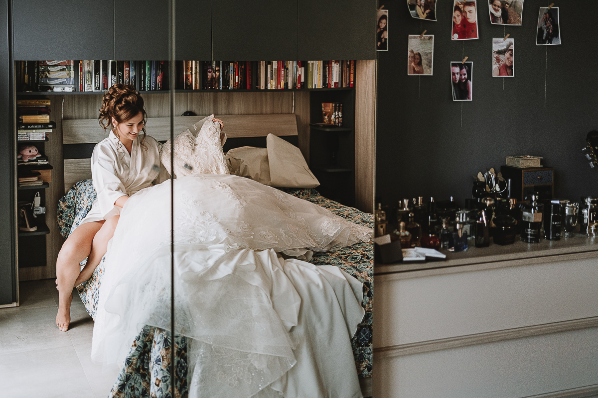Matrimonio Lago di Candia . Preparazione della sposa. Sposa seduta sul letto accarezza il suo abito. riglessa nello specchio dell'armadio.