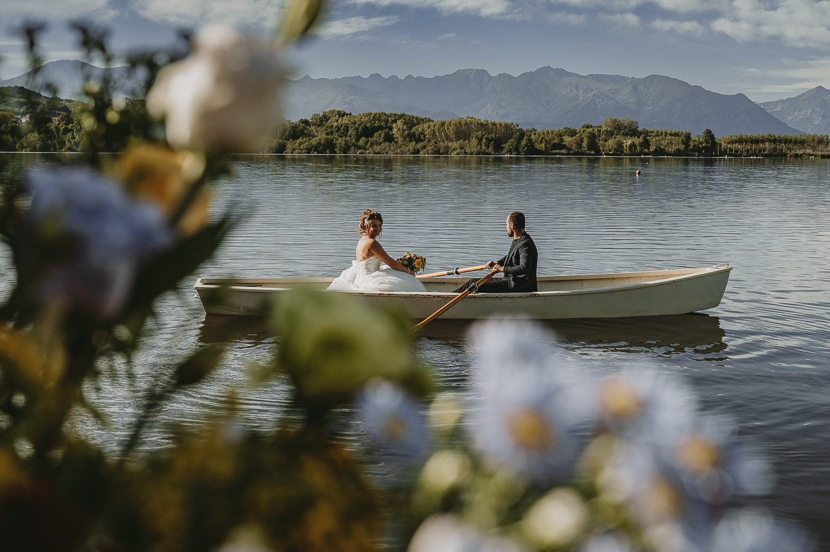 Matrimonio Lago di Candia. Cerimonia. Sposa sulla barca a remi , fotografata dal luogo della cerimonia attraverso i fiori sfocati.