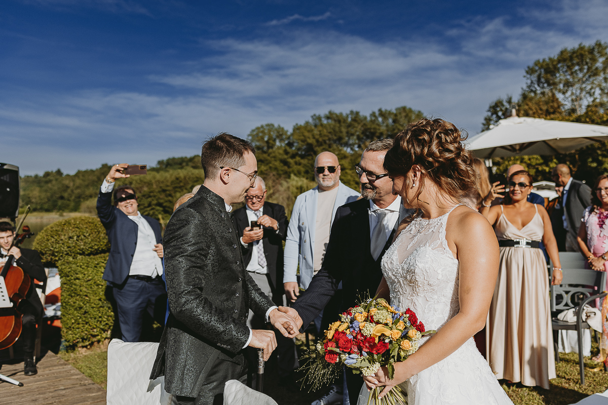 Matrimonio Lago di Candia. Cerimonia. Sposa fa l'ingresso nel luogo della cerimonia sul molo mentre lo sposo aspetta. Il padre saluta lo sposo.